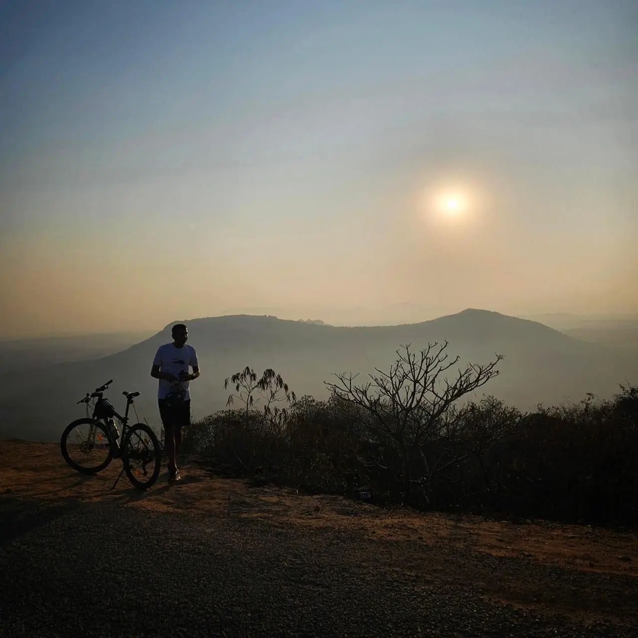 A person standing beside a bicycle on a hilltop at sunset.