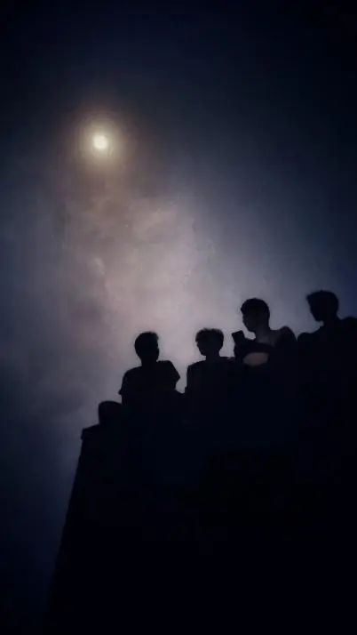 Silhouettes of four friends sitting together beneath a bright moonlit sky.