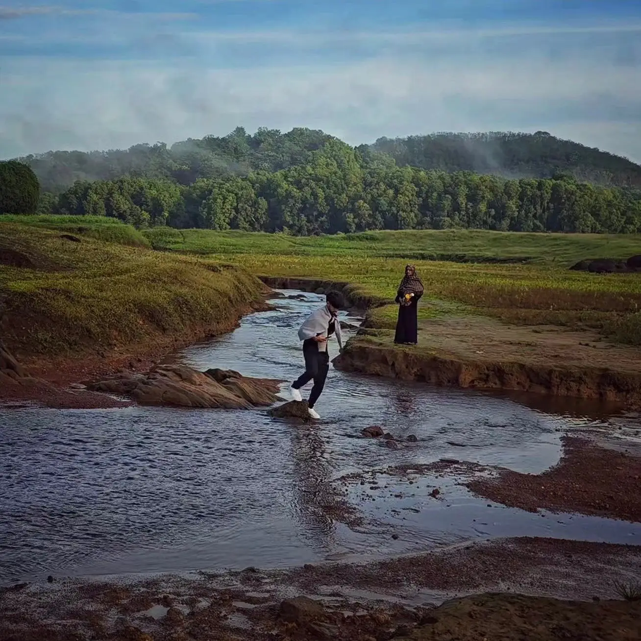 Two people crossing and standing beside a shallow stream in an open grassy landscape.
